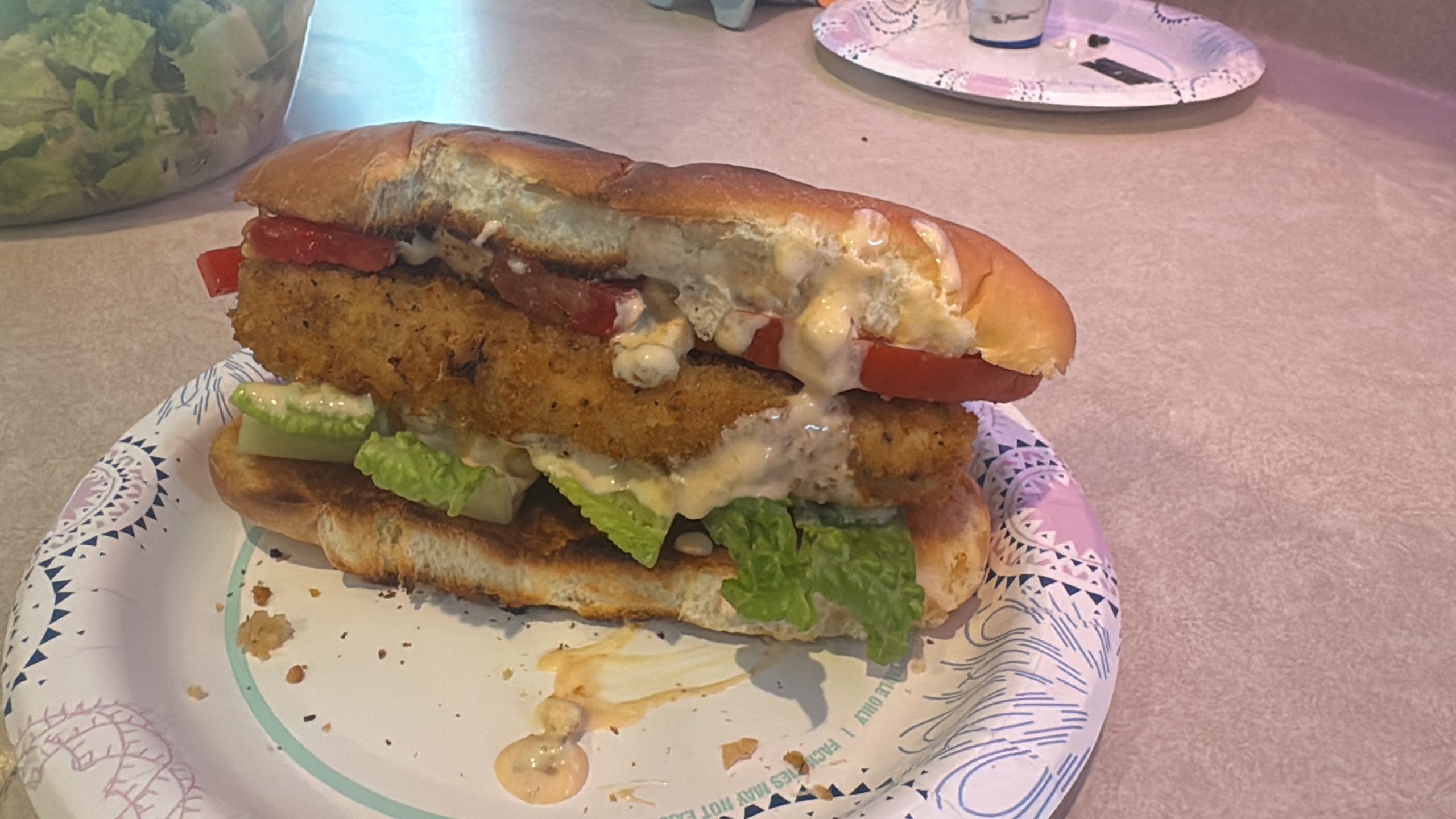 Large leftover fried swordfish sandwich with lettuce, tomato and sauce on a toasted roll.