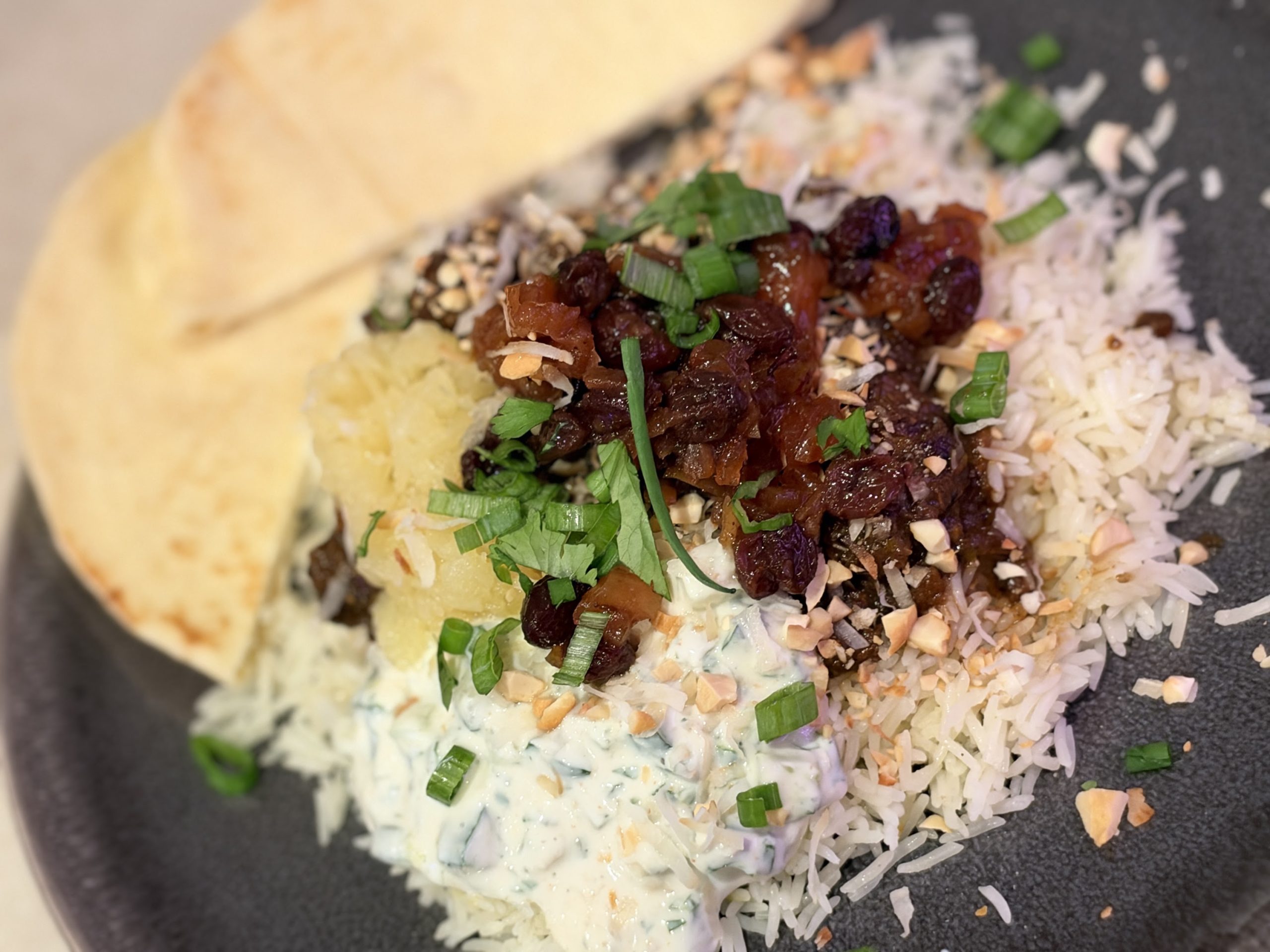 A plate of tender beef curry over basmati rice with chutney, raita, coconut, fresh herbs and naan