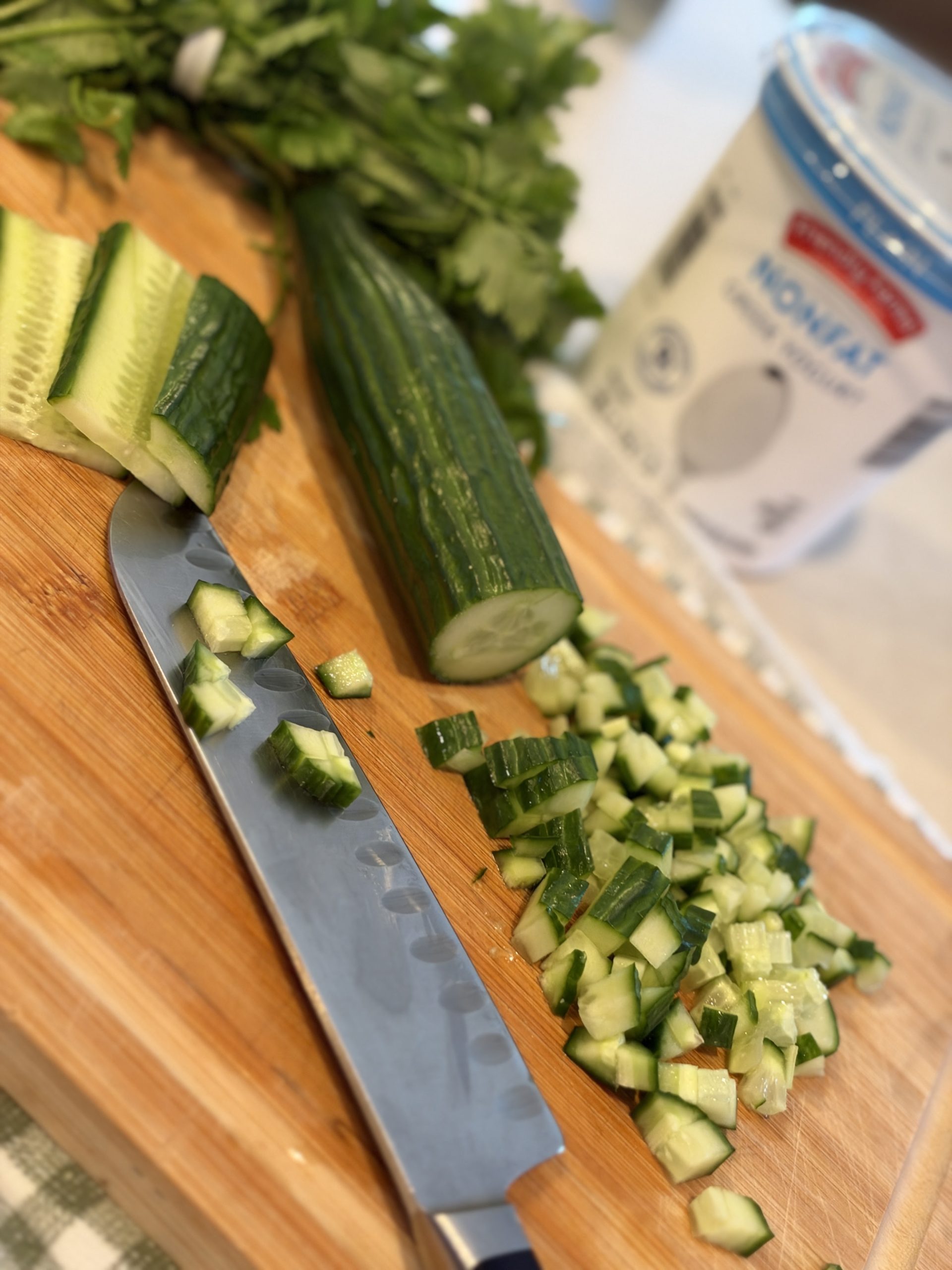 Fresh cucumber being diced beside yogurt and herbs for homemade raita